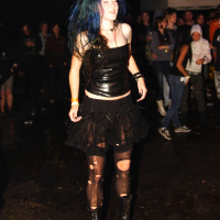 A person with blue hair is dancing outdoors at night during Metalshow 09. They are wearing a black strapless top, a black layered skirt with frayed edges, torn black tights, and black lace-up boots. Other attendees and a white tent are visible in the background. The ground appears wet and muddy.