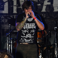 A person with long hair and tattoos on their left arm is performing on stage at a Metalshow, holding a microphone close to their mouth. They are wearing a black t-shirt with white graphics and green shorts. The background features a drum set and a dark banner with partially visible white letters.