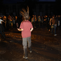 A person with dreadlocks is dancing with their back to the camera at Metalshow '09, a night-time outdoor concert. They are wearing a maroon shirt and dark pants, standing on wet, muddy ground. In the background, a crowd of people watches and mingles. The scene is dimly lit, with a dark building in the distance.