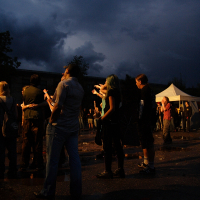 A group of people stands in a poorly lit outdoor area at dusk, facing left. The sky is dark and cloudy. Some gaze upwards, while others chat or look around. A white tent marked "Metalshow '09" is visible in the background on the right. Puddles of water are on the ground, reflecting some of the ambient light.