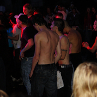 A crowded scene at Metalshow '09, with shirtless individuals enjoying the music. The audience shows a mix of energy levels, with some people raising their hands and others standing still. Colorful stage lights illuminate the crowd, casting vibrant hues of red and orange across the scene.