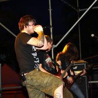 At a 2009 Metalshow, a rock band performs energetically on stage at night. The lead singer, wearing a black T-shirt with graphics and cargo shorts, passionately sings into the microphone. Two guitarists stand behind, one partially obscured. Metal scaffolding and stage lights are visible in the background.