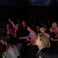 A large crowd of people is dancing and enjoying a nighttime metal show. The group is diverse, with some individuals shirtless and others wearing various clothing styles. The atmosphere appears energized, with people moving actively. Lighting from the stage casts different colors on the attendees, reminiscent of 2009 concerts.