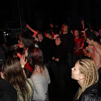A lively crowd is attending a metalshow in a dark, indoor venue. Some are actively dancing and moving, while others are watching the performance. There is a mix of casual and more expressive attire, including one individual with braided hair in the foreground. This event has all the energy of 2009.