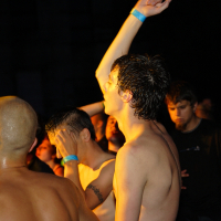 A group of people at a 2009 metal show, illuminated by warm lighting. In the foreground, a shirtless man with wet hair raises his arm energetically. Another man with a shaved head is nearby. The background is filled with people, some facing forward and others turned away.
