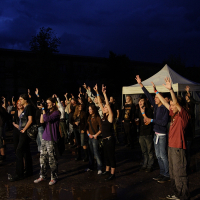 A group of people stands outdoors in front of a white tent at night, raising their hands enthusiastically. The sky is dark and stormy, creating a dramatic atmosphere. Most individuals appear engaged and attentive, suggesting they might be watching a performance at the Metalshow '09.