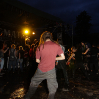 A person with dreadlocks stands in a wet, muddy area at an outdoor concert during nighttime. They wear a red shirt and gray pants. In the background, a crowd of people watches a performance on stage, illuminated by bright yellow light. The sky is dark and cloudy, capturing the essence of the 2009 metal show event.