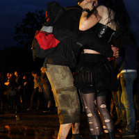 A couple stands closely, embracing each other at a metal event during dusk. The person on the left wears a black cap, black jacket, tan cargo shorts, and a black and red backpack. The other person is dressed in a black top, black skirt, ripped tights, and black knee-high boots. A crowd is visible in the background.