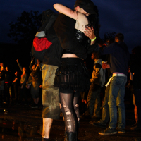 A couple embraces at an outdoor event during nighttime. The woman, dressed in dark clothing with patterned tights and boots, has her arms wrapped around a man wearing a cap, dark jacket, red backpack, and camouflage pants. People in the background of this Metalshow '09 are engaged in various activities.