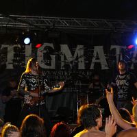 A rock band performs energetically on stage with a large "Stigmata" banner behind them at a 2009 metalshow. The band members wear dark, graphic clothing and play electric guitars and drums. The audience, some with raised hands, enjoys the lively atmosphere under colorful stage lights.