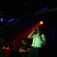 A band performing on a dark stage with dramatic lighting. The lead singer, wearing a white shirt, is holding a microphone and singing passionately at this 2009 metalshow. Two guitarists are on either side, focused on their instruments. In the background, a drum set is partially visible under dim red and blue lights.