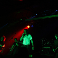 At Metalshow '09, a rock band performs under dramatic red and green lighting. The lead singer, in a white shirt and black pants, stands center stage, singing into the mic. Guitarists flank either side, one in a black shirt. Behind them, the drummer keeps the beat pounding through the night.