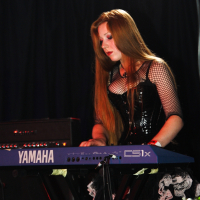 A woman with long red hair is playing a Yamaha CS1x keyboard on stage at Metalshow '09. She is wearing a black corset-style top with mesh sleeves and accessorized with a frilly skirt. A Marshall amplifier is positioned behind her against a backdrop of black curtains. The scene is dimly lit, creating a dramatic atmosphere.