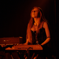 In a 2009 metalshow, a woman with long hair performs on a keyboard under dim, warmly colored stage lighting. Dressed in a black corset, mesh sleeves, and a skirt, she wears a cross necklace. Behind her is a Marshall amplifier. The scene is dark, focusing attention on the performer and her instrument.
