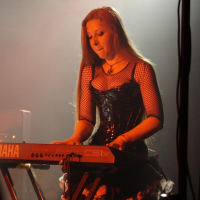 A woman with long hair is playing a Yamaha keyboard on stage at Metalshow '09. She is wearing a black corset dress with fishnet sleeves and a layered lace skirt. The background is smoky and dimly lit, creating a moody atmosphere. A microphone stand is partially visible to the right.