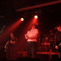 A band performs on a dimly lit stage with red lighting, reminiscent of a 2009 metalshow. The lead singer, in a white shirt, passionately sings into the mic, head thrown back. Two guitarists and a bassist stand nearby, playing their instruments. The drummer is in the background behind the drum kit, partially visible.
