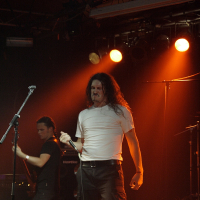 A long-haired man in a white shirt and dark pants passionately sings into a microphone on a dimly lit stage with orange lighting at Metalshow '09. Another musician in black stands behind him, playing a guitar. Music equipment and spotlights are visible in the background, adding to the intense atmosphere.