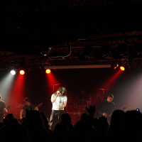 A rock band performing on stage with five members visible. The lead vocalist is in the center, singing passionately into a microphone. Other band members are playing guitars, a keyboard, and drums. The stage is lit with vibrant red and white lights, reminiscent of a 2009 metalshow, and an audience is silhouetted in the foreground.