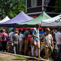 A bustling outdoor market with numerous people interacting under colorful tents at Kalnciema kvartāla. Some individuals are browsing, while others are engaged in conversation. The green, purple, blue, and white tents add to the vibrant scene of modes diena set against a backdrop of trees and buildings.