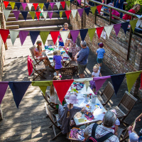 A vibrant outdoor market scene at Kalnciema kvartāla with people seated at wooden tables under colorful string bunting. The area is divided into upper and lower levels with brick walls. Patrons, including adults and children, are engaged in conversations and activities surrounded by festival tents and greenery on this lively modes diena.
