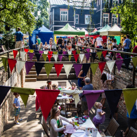 A lively outdoor gathering in the Kalnciema kvartāla garden features colorful triangular bunting strung along fences. People are seated at wooden tables, engaging in various modes of activities. Families with children and various stalls or tents are in the background. Trees and brick walls frame the cheerful atmosphere.