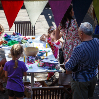 A group of children and an elderly man gather around a table outdoors at Kalnciema kvartāla, engaging in arts and crafts. The children, seated on wooden chairs, are cutting and gluing colorful paper. Triangular fabric banners hang overhead, adding a festive atmosphere to this lively modes diena event. Several other adults stand nearby observing.