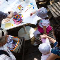 Top-down view of a family at a wooden table engaged in arts and crafts during the Kalnciema kvartāla modes diena. A child in a striped hat and pink pants draws on paper, while another child in a white hat is held by an adult. Various art supplies are scattered on the table as observers in white and plaid shirts watch.