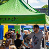 A bustling outdoor market scene at Kalnciema kvartāls with people gathered around a green tent labeled "EcoCatering.lv." The crowd includes diverse individuals, some browsing items, while a man in a gray T-shirt and cap stands in the foreground, and a woman with a flower crown looks at a stall on this vibrant modes diena.