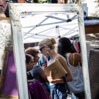 A large, ornate, white-framed mirror reflects a woman in a blue dress holding a brown folder at an outdoor market. Behind the mirror at Kalnciema kvartāls, several people browse items under a white canopy, and a woman with a red flower in her hair stands behind a wooden counter. Bunting is visible in the background.