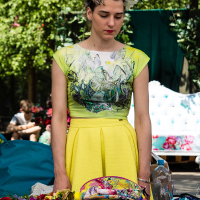 A person with a floral headpiece stands behind a table displaying various colorful accessories, including flowers, purses, and hats. They are wearing a bright yellow skirt and an artistic green and white top. The backdrop of Kalnciema kvartāla features green foliage and people sitting on benches, celebrating modes diena.