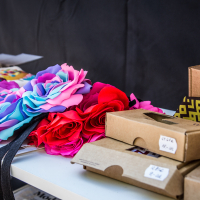 A table in Kalnciema kvartāls features colorful artificial flowers in shades of pink, purple, and blue, beside a stack of cardboard boxes and patterned fabric. The boxes have clear windows and small handwritten labels. The background is blurred, focusing on the charming montage of items on the table.
