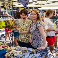 Two women, one with short brown hair and the other with long blonde hair, stand at a vibrant outdoor market stall in Kalnciema kvartāla. Both wear patterned tops. The stall displays colorful items like clothes and textiles. More people are present in the busy background, along with a painting on an easel.