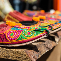 Close-up view of intricately designed traditional footwear, featuring vibrant red fabric with colorful thread embroidery in swirling patterns. The shoes, showcased at Kalnciema kvartāla modes diena, are displayed on a wooden surface with a blurred background indicating an outdoor market setting.