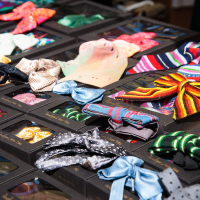 A display table at Kalnciema kvartāls is filled with variously colored and patterned bow ties, neatly arranged in black boxes with clear plastic windows. The ties have designs ranging from polka dots and stripes to solid colors in bright and pastel hues, perfect for modes diena in Latvija.