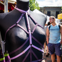 A mannequin dressed in an intricate black chest harness with purple straps is displayed outdoors on a sunny day during Kalnciema kvartāla modes diena. Surrounded by market stalls with colorful tents, a man in a light blue tank top walks by in the background, against a backdrop of trees and a partially cloudy sky.