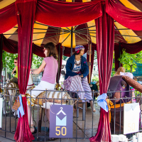 A colorful carousel with red curtains at the Kalnciema kvartāla features adults riding decorated carousel horses. One rider, wearing a hat and striped pants, is gazing to the side, while another is facing forward. The carousel, celebrating modes diena, is adorned with blue banners displaying the number 50. Trees and a picket fence are visible in the background.