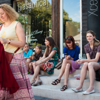 A woman with curly hair, wearing a white t-shirt and patterned skirt, holds a red garment while walking. Five people are sitting on stone steps: three children, an older woman in a floral dress, and a young woman in a purple top and skirt. They appear relaxed outside the Kalnciema kvartāla storefront with large windows on this modes diena.