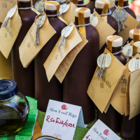 A display of dark glass bottles with vintage-style labels and silver wax seals is shown at Kalnciema kvartāla. Each bottle has a brown paper tag attached with twine. The sign in front reads, "Slow Food Riga" and displays a price of 8.50 EUR. A small jar of dark liquid sits on a green tablecloth, capturing the essence of modes diena.