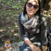 A woman wearing a gray dress and large sunglasses is sitting outside on a stone bench in Kalnciema kvartāla. She has a white and gray patterned scarf around her neck, holding a smartphone in one hand. A stemmed glass with a light-colored beverage rests beside her as dappled sunlight filters through the trees, capturing the essence of modes diena.