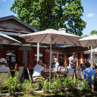 People seated at wooden tables under large patio umbrellas, enjoying meals and conversations at an outdoor cafe. The scene is vibrant, full of social activity. A rustic building with red-trimmed windows in Kalnciema kvartāla and lush green trees in the background complete the lively, sunny ambiance on this modes diena.