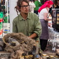 A vendor with long hair and glasses wears a green shirt and stands behind a market stall at the vibrant Kalnciema kvartāla. The stall displays jars of preserves, bottles of liquids, and a variety of handmade goods. Other people are visible in the background, including a woman in a pink headscarf, under white canopies.
