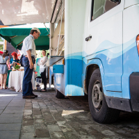 A food truck with a white and blue color scheme is parked on the street at Kalnciema kvartāls. Customers, including a man in a white shirt and another in jeans and a cap, stand nearby. In the background, people gathered under a green canopy near tables with various items enjoy the sunny modes diena. The pavement is a mix of bricks and tiles.