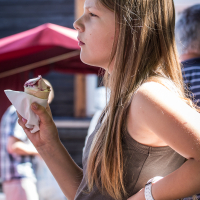 A young girl with long brown hair, wearing a gray sleeveless top and a white wristwatch, stands outdoors holding an ice cream cone in her left hand. She looks to the left with a thoughtful expression. Behind her, during modes diena at Kalnciema kvartāls in Rīga, there are people, a red umbrella, and a building with dark siding.