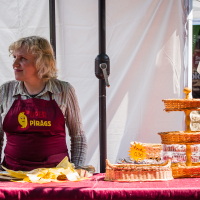 A person with light-colored, curly hair wearing a striped shirt and a red apron labeled "Rūder Pīrāgs" stands behind a booth at the Kalnciema kvartāla market. They are next to a tiered wicker stand holding baked goods. The booth has a maroon tablecloth, with yellow napkins and pastries on the table.