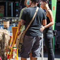 Three people, two men and one woman, stand closely together at an outdoor market stall during Kalnciema kvartāla modes diena. The woman, on the right, wears black clothing, and one man, with dreadlocks and cargo shorts, carries a shoulder bag. The other man is partially visible. Market tents and various products fill the background.