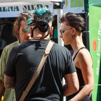 Three people are standing at Kalnciema kvartāls, engaged in conversation. One person has green highlights in their hair and wears a light green shirt. Another has dreadlocks and wears a black shirt with a shoulder bag strap. A third person with dark, short, side-shaved hair wears a black tank top.