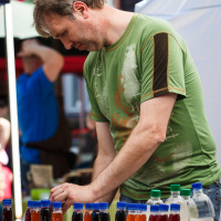 A vendor wearing a green T-shirt is standing at an outdoor market stall in Kalnciema kvartāls, arranging various colored bottles of juice or syrup. The bottles are neatly lined up on the table, each labeled with different flavors. The backdrop captures other market stalls and blurred people enjoying modes diena.