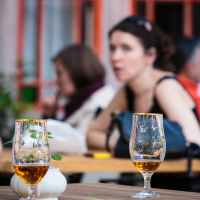 A close-up image of two half-full stemmed beer glasses on a wooden table outdoors at Kalnciema kvartāla's modes diena. In the background, there are two people, one blurred, seated at another table. The scene appears to be at a café or beer garden, with soft focus on greenery and red window frames in the background.