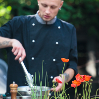 A chef with a shaved head, wearing a black double-breasted chef's coat, and holding tongs is focused on cooking at an outdoor setting during Kalnciema kvartāla modes diena. In the foreground, vibrant red poppies and green foliage are slightly out of focus. The background is a blur of green trees.