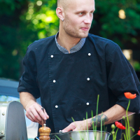 A chef with a shaved head and earrings, wearing a black double-breasted chef's coat, is standing outdoors at Kalnciema kvartāls preparing food. He holds a metal bowl and stirs its contents with a spoon, while a wooden pepper mill rests on the countertop beside him. Lush green trees are in the background.